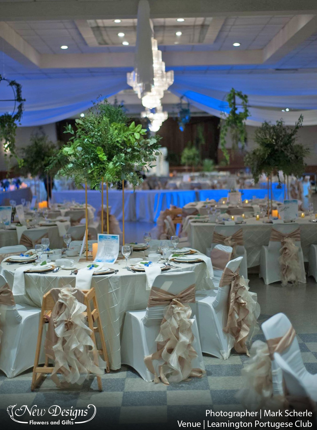 A tall centerpiece on a 4 posted metal frame, topped with a wild greenery arrangement, in a hall of crisp white linen with trailing textured tufts of nude material swirling down the back of each chair.